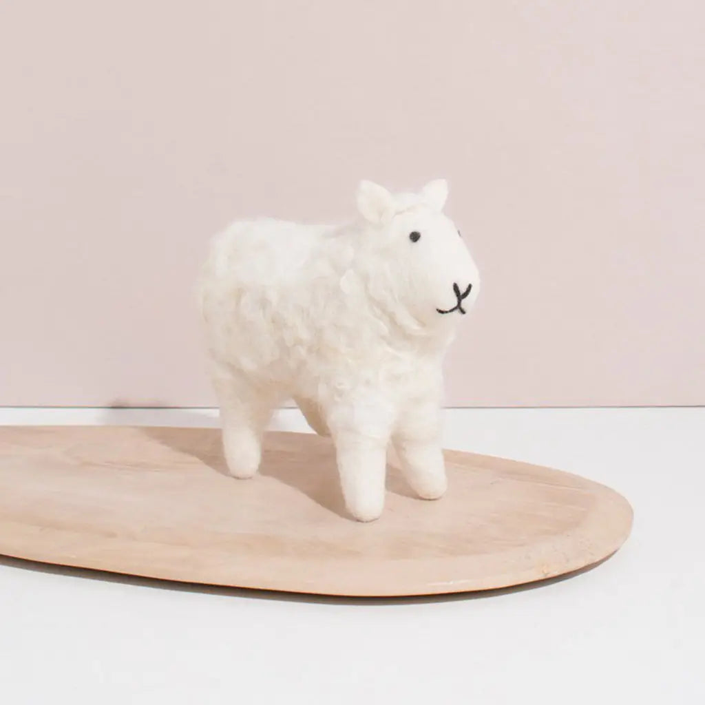 A small, fluffy Hand Felted White Wool Sheep figurine, handcrafted from naturally-dyed wool with a simple, smiling face stands on a light wooden oval base against a soft pink background.