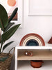 A minimalist interior featuring a stylish Handmade Rainbow Stacker - Terracotta on a white shelf, with a potted plant nearby. Below, a wooden craft object and a terracotta bowl add warmth to the space.