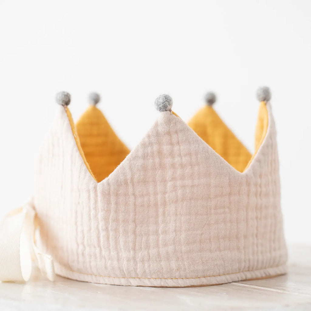 A soft, handmade Reversible Crown in plush, light beige organic cotton with golden yellow peaks topped with gray pom-poms, displayed against a light background.