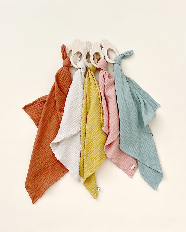 A collection of colorful Organic Muslin Lovey with handles, neatly arranged in a fan shape on a light beige background. Colors include orange, red, yellow, green, and blue. Each bag is handmade in