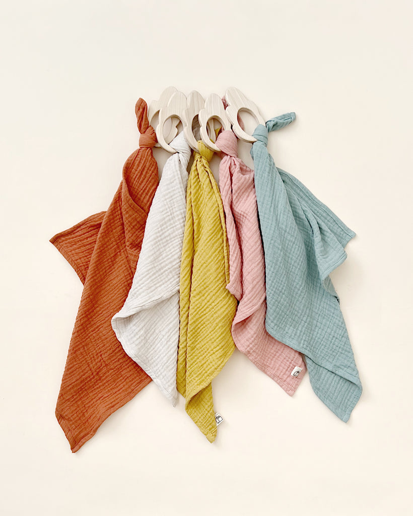 A collection of colorful Organic Muslin Lovey with handles, neatly arranged in a fan shape on a light beige background. Colors include orange, red, yellow, green, and blue. Each bag is handmade in