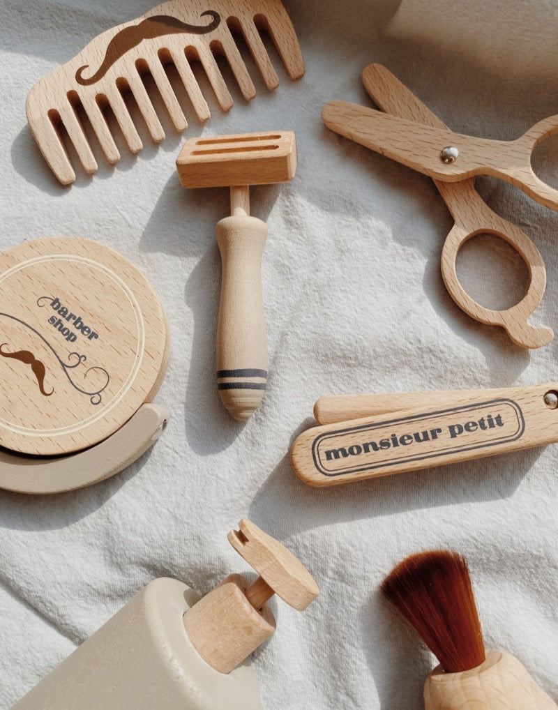 Various Pretend Play Wooden Barber Set tools, including combs, scissors, and brushes, laid out on a textured light fabric background. Some items are branded with names.
