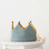 A reversible Reversible Crown handmade from organic cotton in teal and mustard colors with small pompoms on its peaks, placed on a white stool against a plain background.