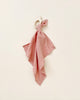 A soft pink Organic Muslin Lovey tied to a wooden teether, displayed against a light beige background. The cloth is draped elegantly with a neat bow at the top.