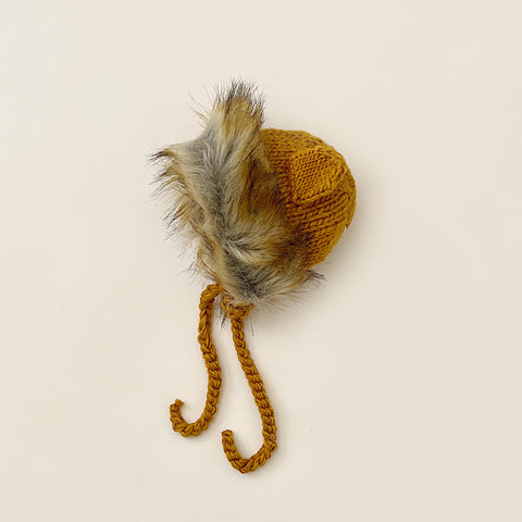A small, hand-knit acrylic hat with a fluffy faux fur pom-pom on top and braided ties, displayed on a light beige background, The Blueberry Hill Lion Hat.