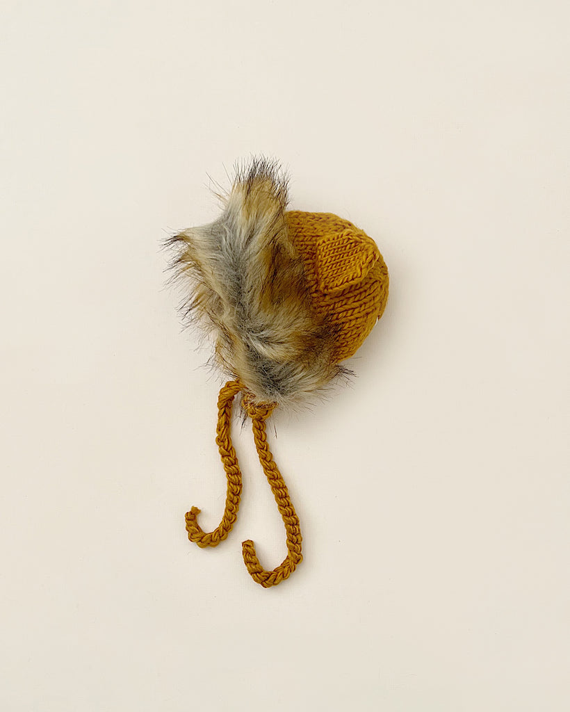 A small, hand-knit acrylic hat with a fluffy faux fur pom-pom on top and braided ties, displayed on a light beige background, The Blueberry Hill Lion Hat.