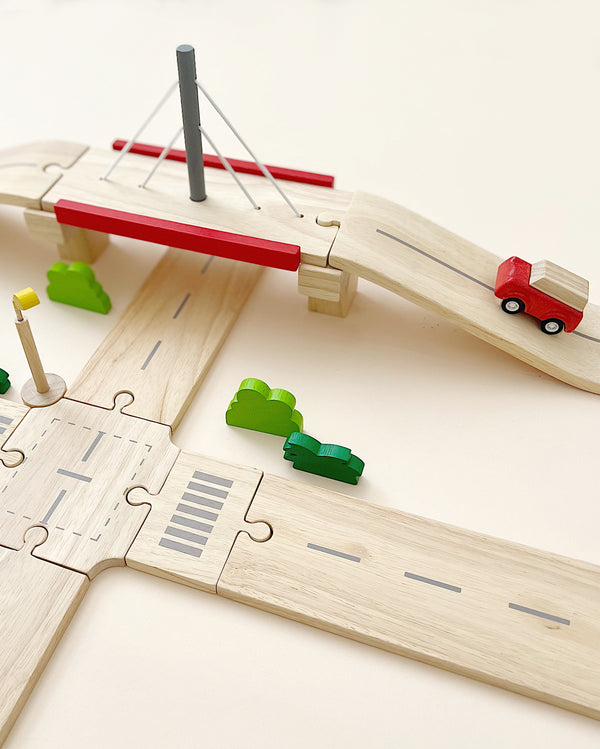 A Wooden Road System Deluxe set depicting a roadways scene with a bridge, a red toy car, three green bushes, and traffic signs on a light beige background.