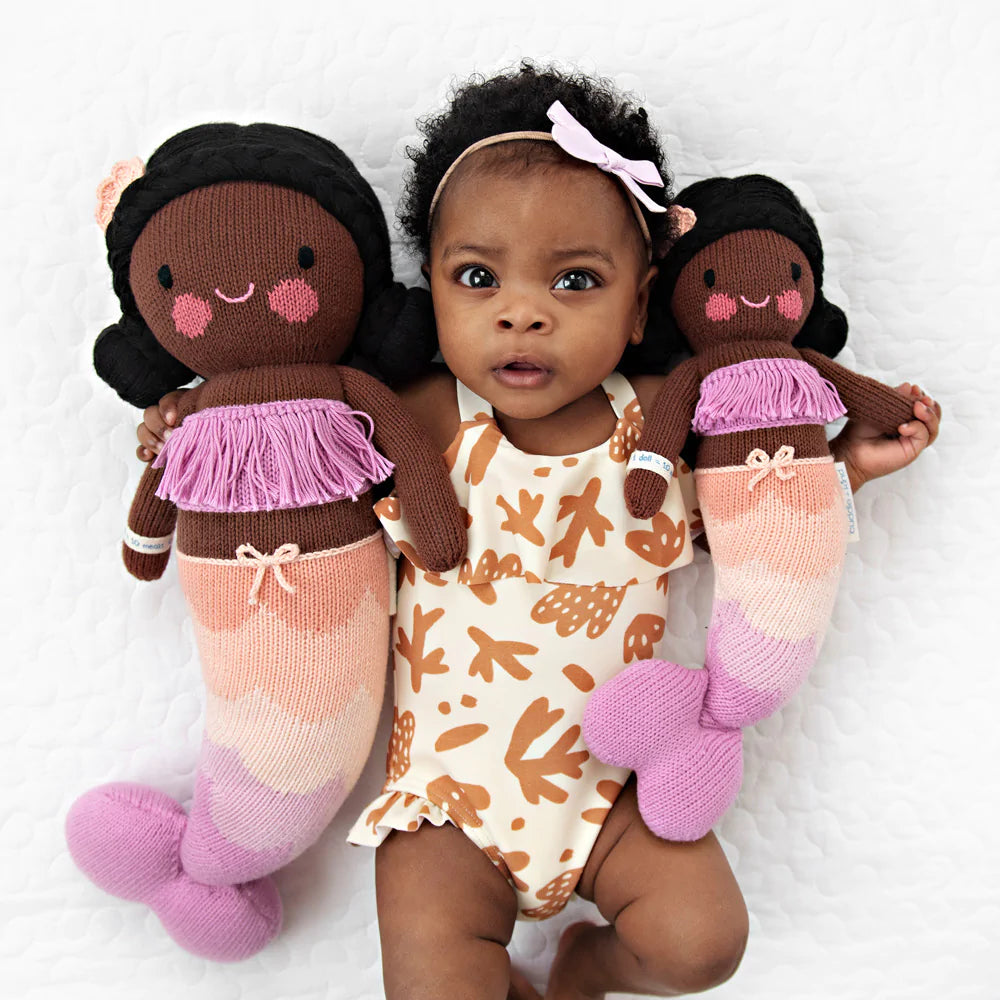 A baby with a light pink bow in her hair lying on a white background, flanked by two Cuddle + Kind Maya the Mermaid plush dolls with brown and pink details, filled with hypoallergenic polyfill, wearing a pattern.