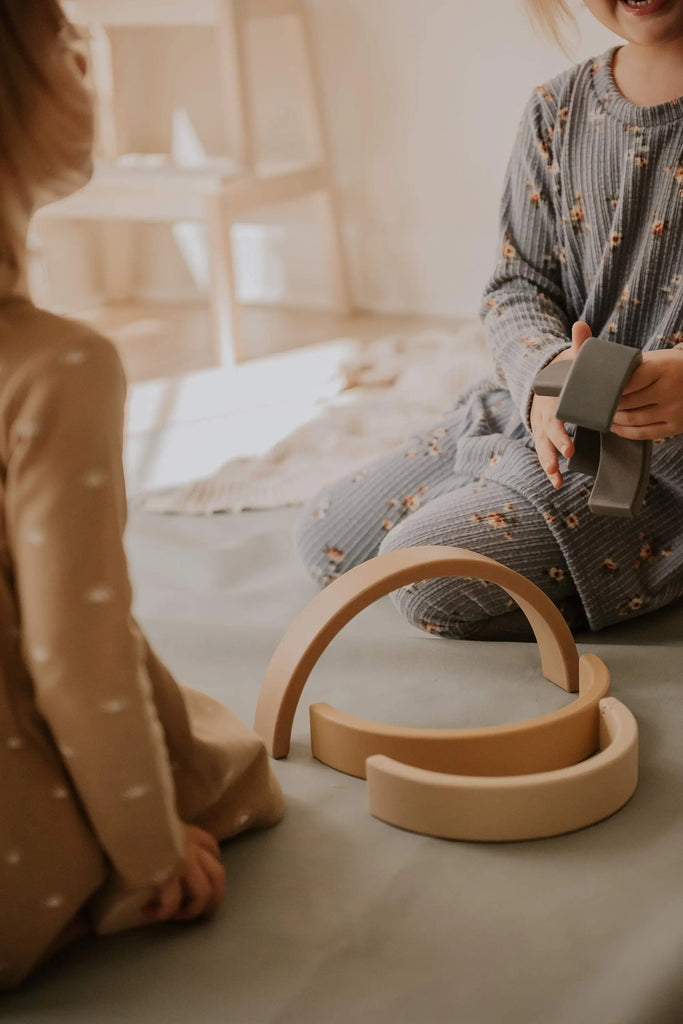 Two children are sitting on the floor, playing with the **Handmade Rainbow Stacker - Mustard**, a non-toxic stacking toy made of wooden arches and blocks. One child is holding a couple of pieces, while the other watches. Both are dressed in soft, neutral-toned pajamas with patterns. The background is softly lit with warm, natural light.