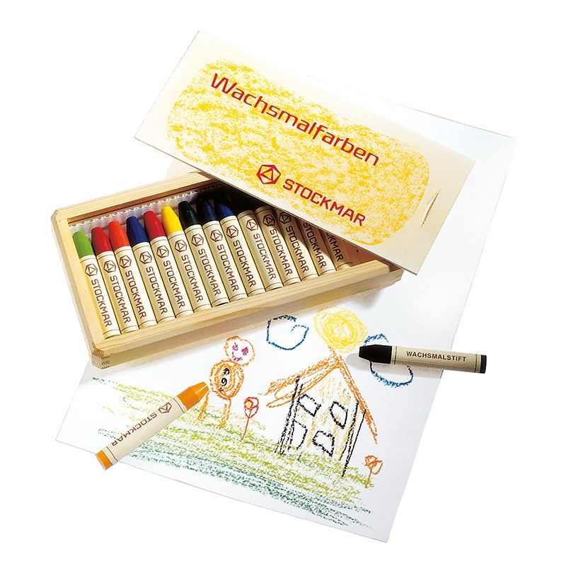 A box of Stockmar Wax Stick Crayons Wooden Box - 16 Assorted is open next to a child's crayon drawing of a house and sun, with a loose crayon and wrapper in the foreground.