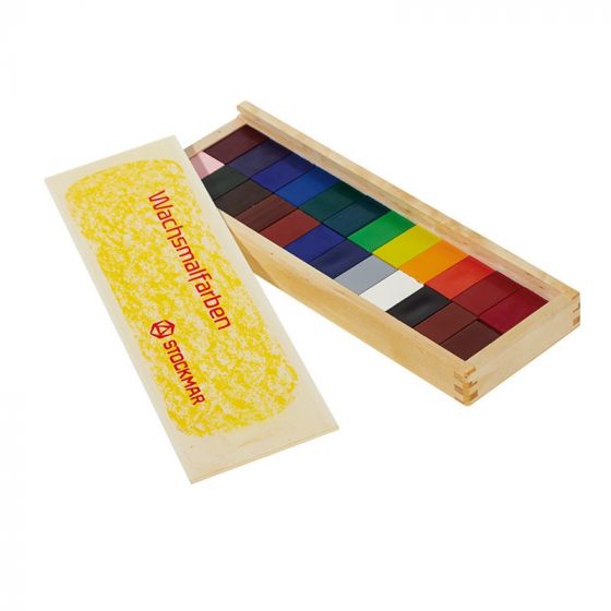 A wooden box containing a set of colorful Stockmar Wax Block Crayons for children, each wrapped in paper, arranged next to a yellow block placed on its lid.