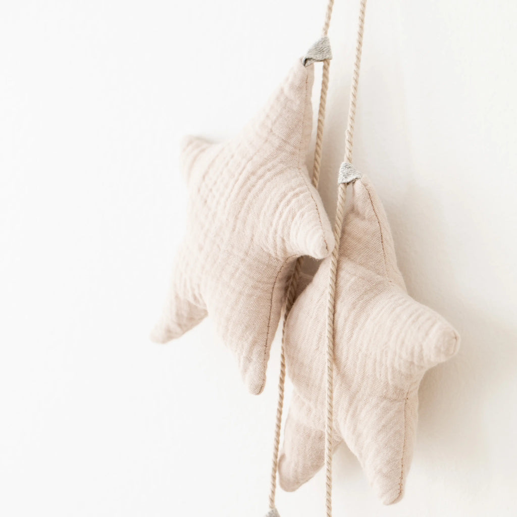 Two beige organic cotton Handmade Star Garland ornaments hanging on a white wall, linked by a light grey cord, in a clean, minimalist setting.