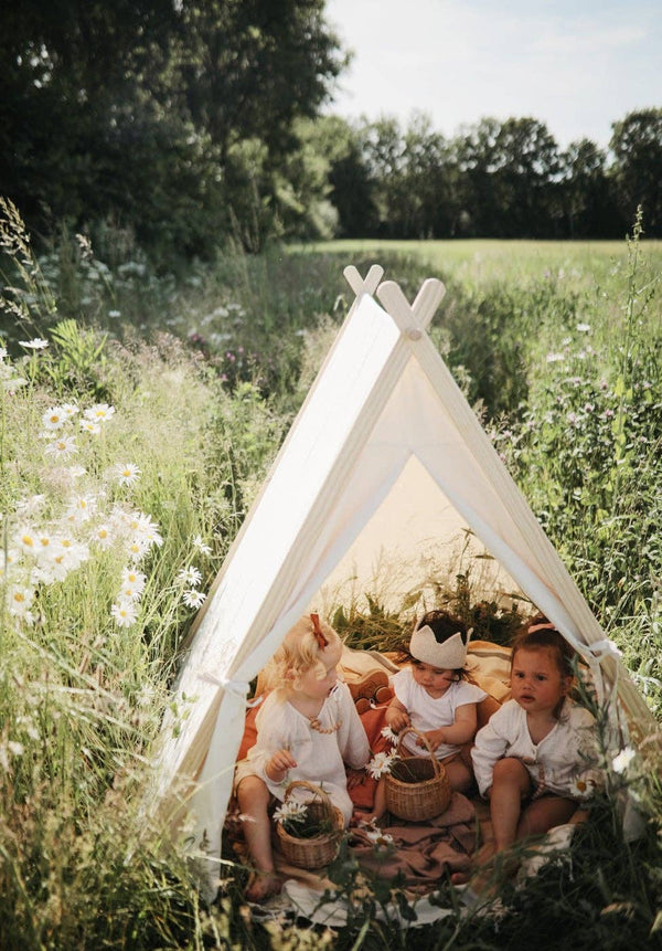 Three young children inside the Fisher-Price Kid's Indoor/Outdoor Play Tent in a lush meadow, surrounded by wildflowers under a sunny sky. They are dressed in light outfits, sharing a relaxed picnic moment.