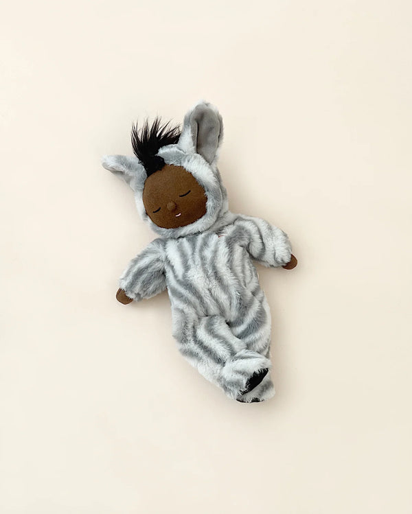 A soft toy depicting a brown-skinned collectible Olli Ella Zebra Mini Doll with black hair, dressed in a plush onesie zebra costume, lying on a light beige background.