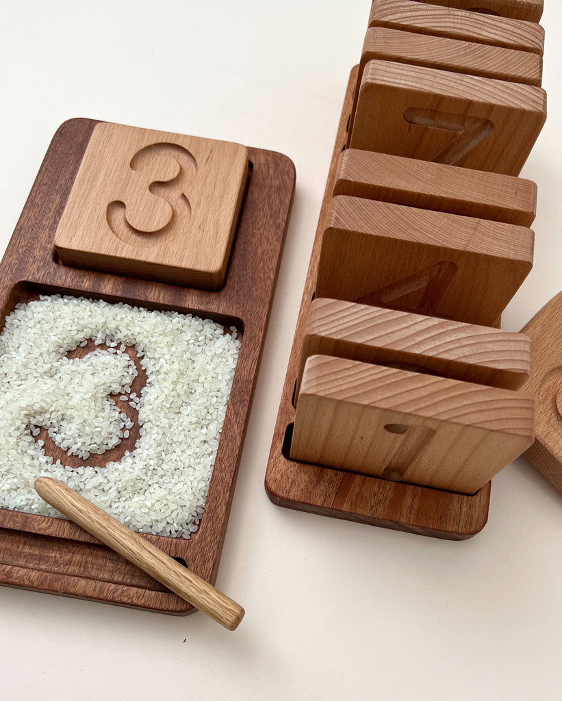 The Wooden Counting & Tracing Tray - Made in USA holds rice and a pen beside number tiles. The number 3 is traced in the rice and displayed on a tile, with other numbers stacked nearby—perfect for hands-on counting and tracing practice.