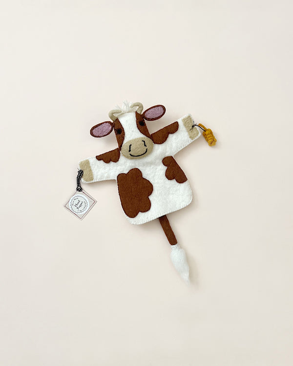 A hand puppet cow with a whimsical design, needle felted from organic wool, featuring brown patches and a small bell on its tail, set against a light beige background.
