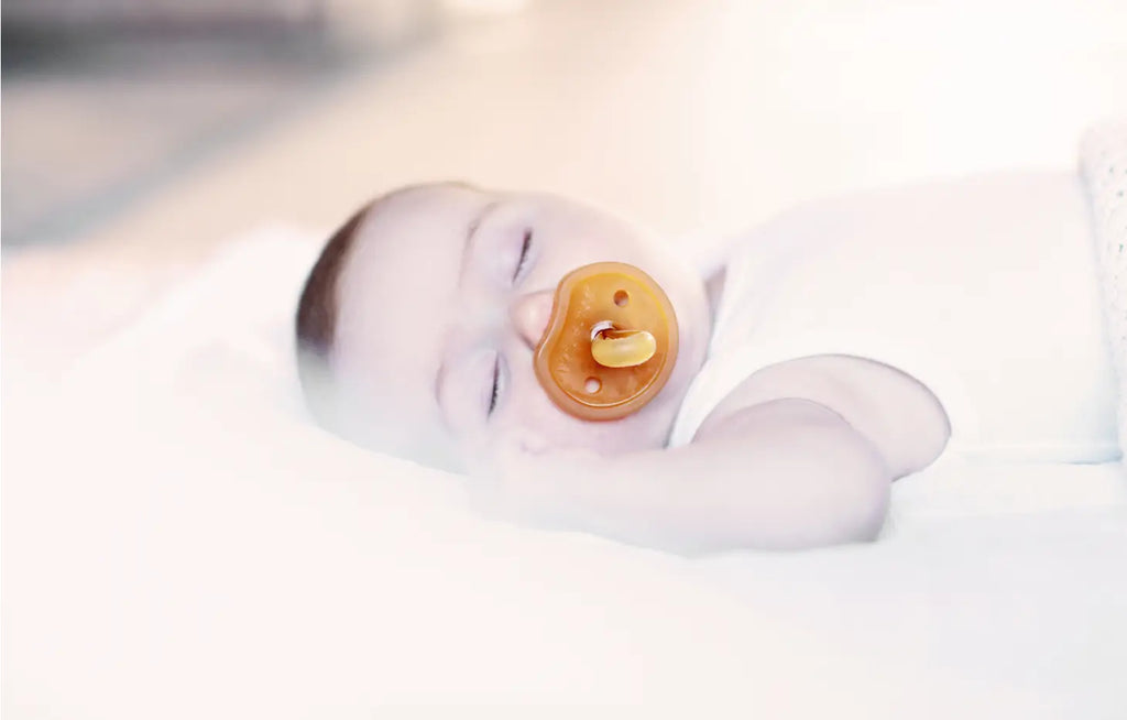 A peaceful newborn with soft skin sleeping soundly on a white blanket, using a Natursutten Butterfly Pacifier | 0-6 Months, small. The ambiance is soft and serene with a gentle light.
