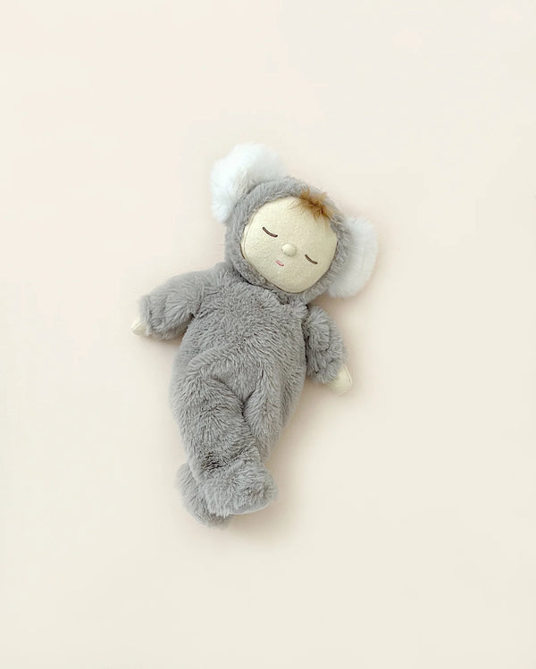 A soft plush toy resembling a sleeping child in a cozy Olli Ella Koala Moppet Doll suit with white ears, lying on a light beige background.
