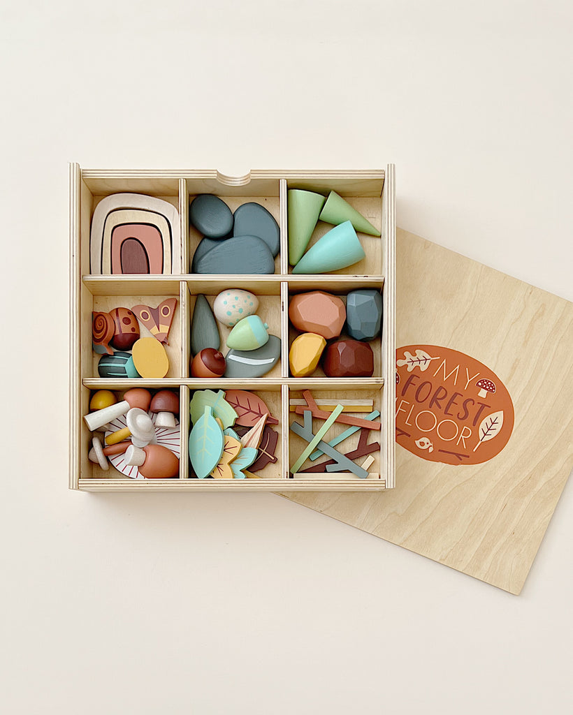A wooden tinker tray containing various colorful, abstract wooden shapes and pieces, arranged in compartments, next to a lid with "My Forest Floor" etched on it, placed on a cream background.
