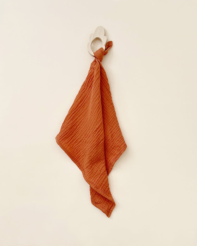 A burnt orange **Organic Muslin Lovey** creatively knotted and hanging on a white hook against a plain, light-colored background.