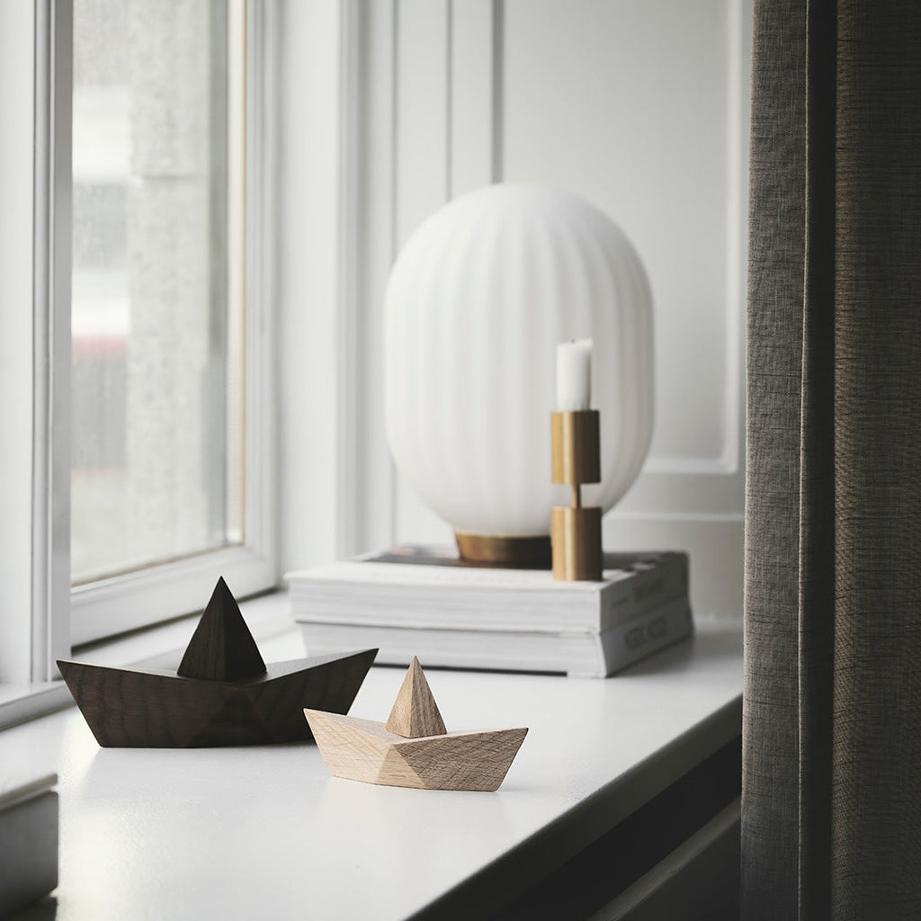 Two Boyhood Admiral, Small Oak origami boats, one dark and one light, on a windowsill beside a round white lamp that sits on books, with a view of an overcast day outside. One of the boats