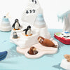 A whimsical display featuring miniature animal figurines, including penguins and seals, staged on a stylized ice and water set crafted from sustainably sourced wood with small boats floating nearby, the Wooden Antarctica Music Box.