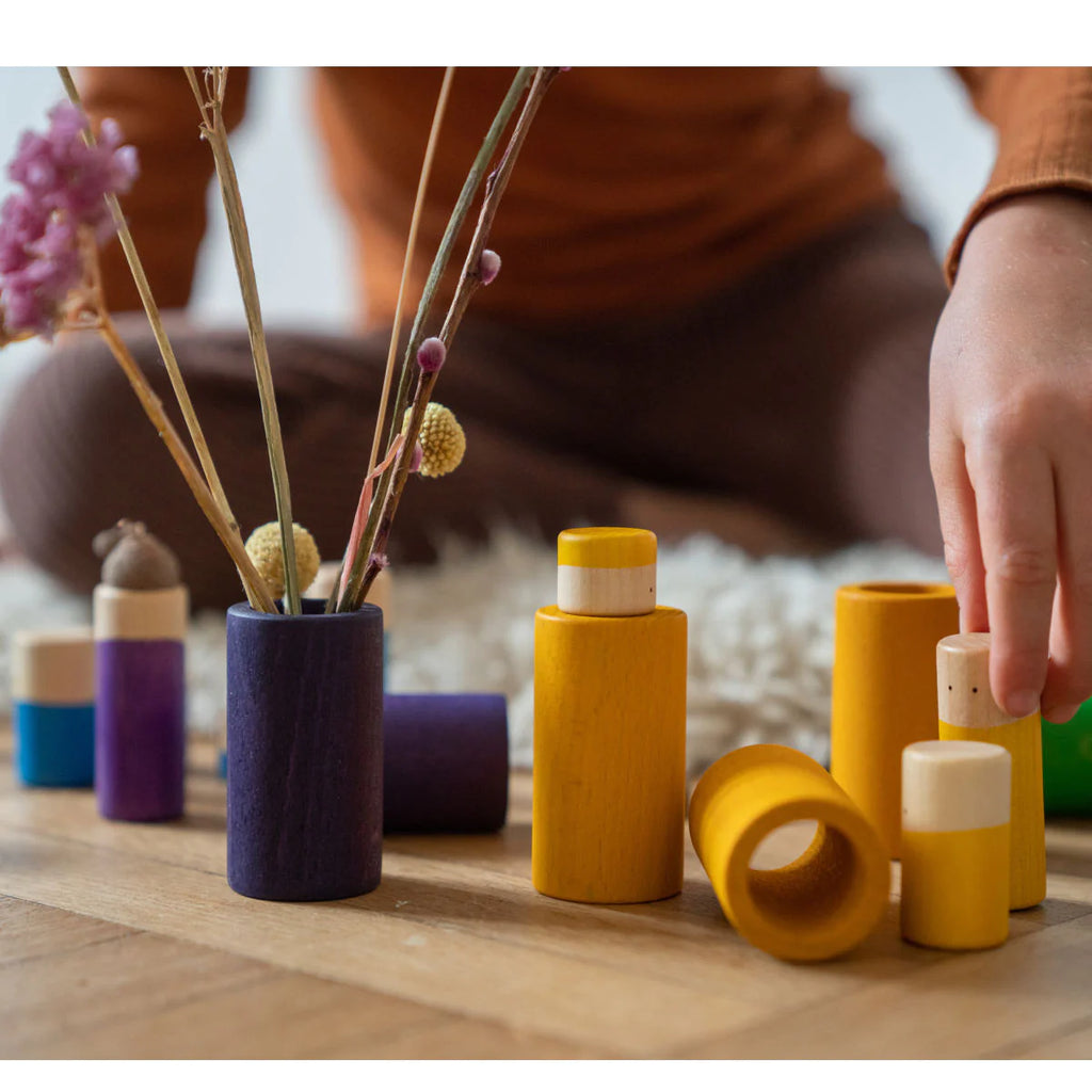 A close-up of a person's hands arranging Grapat LA Non Basic Colors play set 36 pcs on the floor, with a vase of dried flowers in the background.