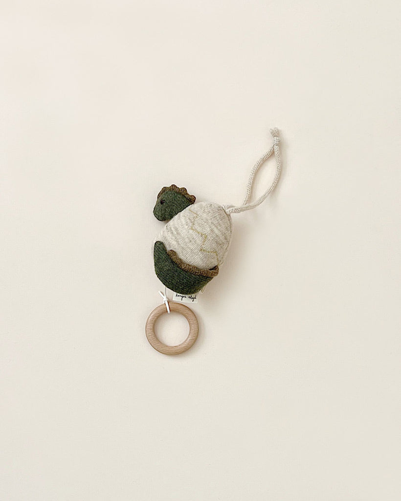 A soft, plush dinosaur toy with green and white lambswool fabric hangs from a white string. Attached to the bottom of the Lullaby Activity Pull Toy - Dinosaur is a wooden ring. The toy's simple design is set against a plain, light-colored background.