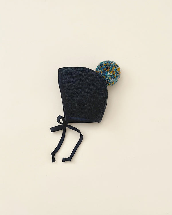 A small, dark blue denim Briar Baby Eon Pom bonnet with black ties and a colorful yarn pom-pom attached, handmade in the USA, displayed against a plain light background.