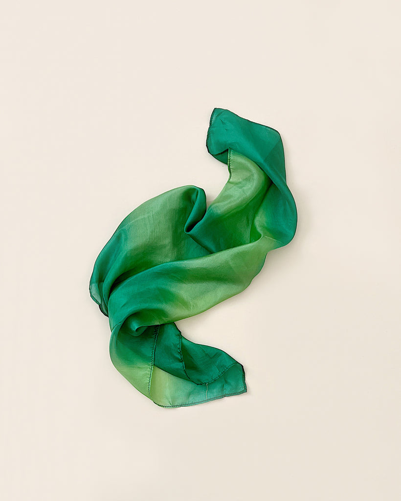 A green Playsilk scarf with gradations of light and dark shades, elegantly draped in a loose, flowing manner on a light beige background.