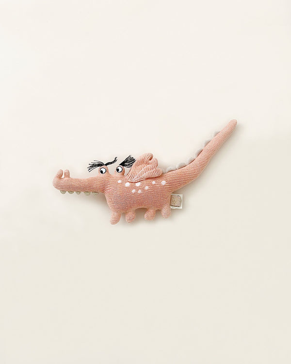 A Baby Crocodile Rattle in the shape of a pink crocodile with a textured body, wearing a playful expression and positioned against a light background. This soft cushion is made from 100% cotton.