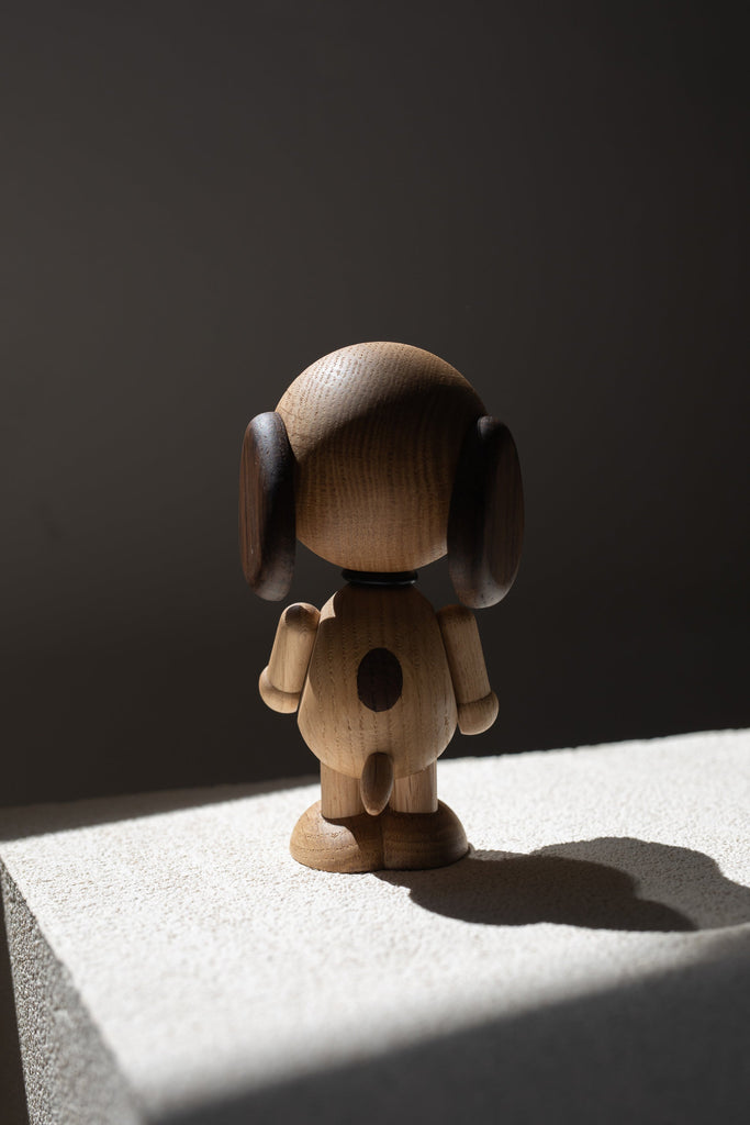 A wooden figurine of Boyhood Snoopy, small with oversized headphones stands on a stone surface, dramatically lit by natural light casting strong shadows.