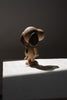 A wooden figurine of Boyhood Snoopy, Small standing on a textured surface, lit by sunlight creating a defined shadow. The figure has a rounded shape and smooth, natural wood tones.