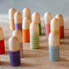 A collection of Grapat Buds/Brots, small wooden figures painted with colorful striped patterns standing on a wooden surface, each figure has a simple, smiling face.