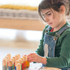A young girl wearing a green jumper is intently arranging colorful Grapat Buds/Brots from sustainable forests on a table, symbolizing learning and concentration.