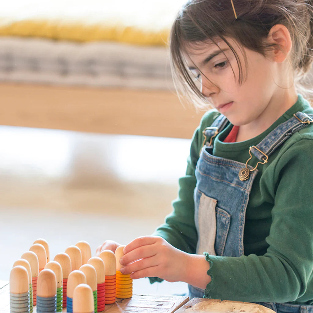 A young girl wearing a green jumper is intently arranging colorful Grapat Buds/Brots from sustainable forests on a table, symbolizing learning and concentration.