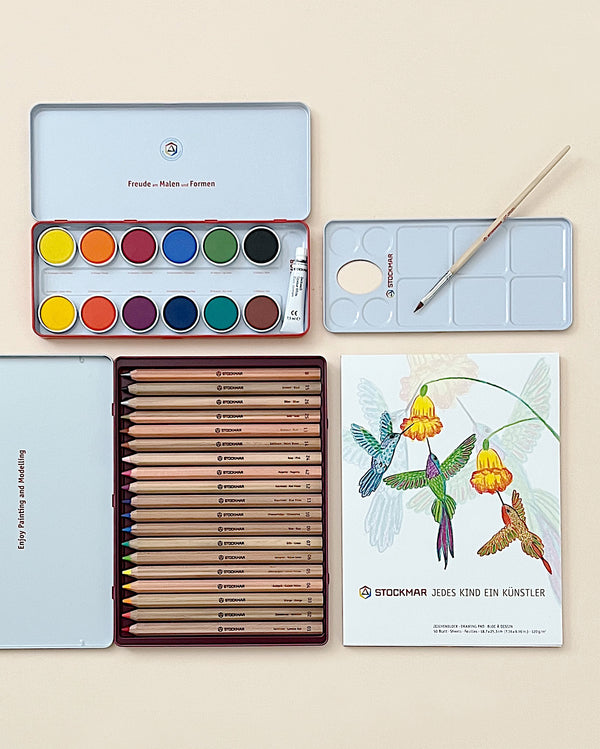 An assortment of art supplies including opaque watercolors, brushes, hexagonal colored pencils, and Stockmar drawing pads displayed on a pale surface. The supplies are neatly organized, blending creativity with order.