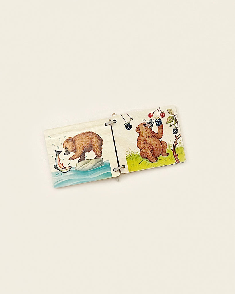 Two wooden picture book pieces made from sustainably harvested trees, connected together, featuring illustrations of a bear fishing in the river and another bear on a grassy knoll with berries and bees around it, set against
