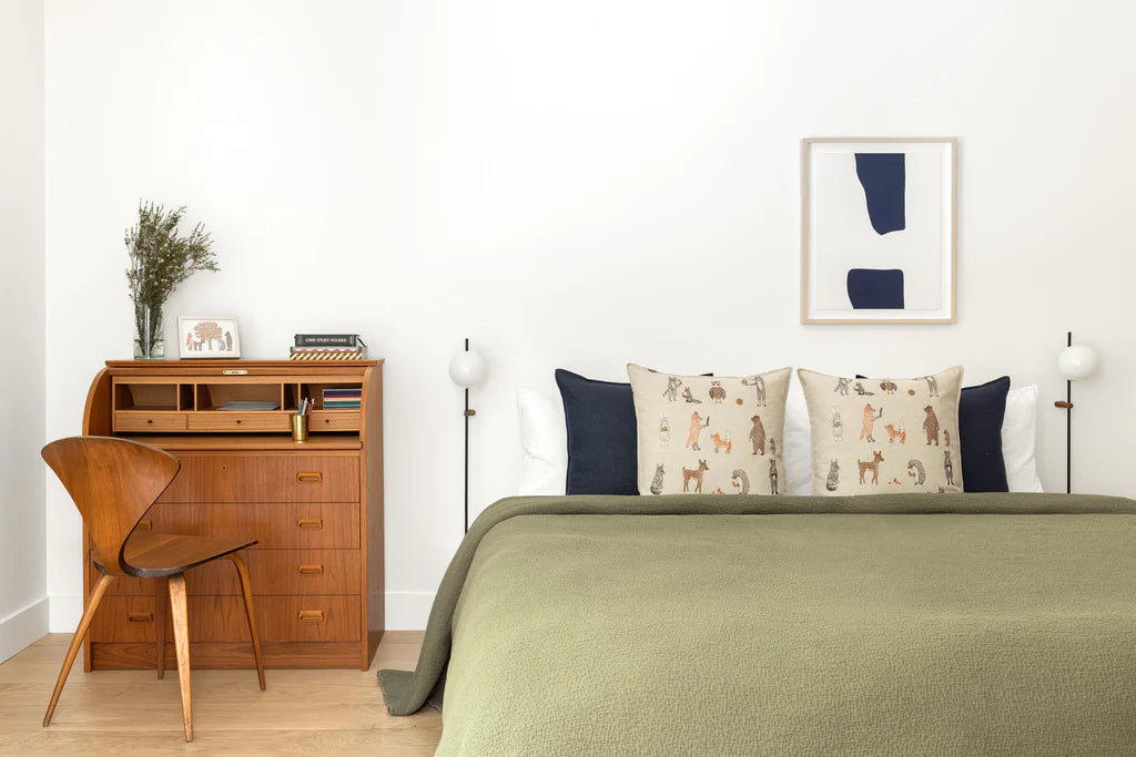 A minimalist bedroom featuring a green bedspread, a wooden desk with shelves, a classic chair, and a framed abstract painting on the wall. Two Coral & Tusk Woodland Friends Pillows with animal prints and white bed lamps are visible.