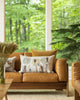 A cozy living room corner with a caramel-colored leather couch adorned with Coral & Tusk Orchard Pocket Pillows. Large windows offer a view of a lush green forest. A potted plant and dried flowers enhance the tranquil ambiance.