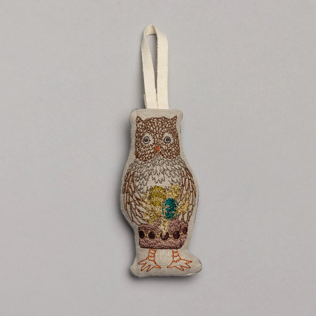 The Coral & Tusk Owl Ornament, with intricate embroidery in brown, gold, and green threads, brings a festive forest theme to life. It hangs from a beige ribbon against a plain gray background, perfect for adding charm to your Christmas collection.