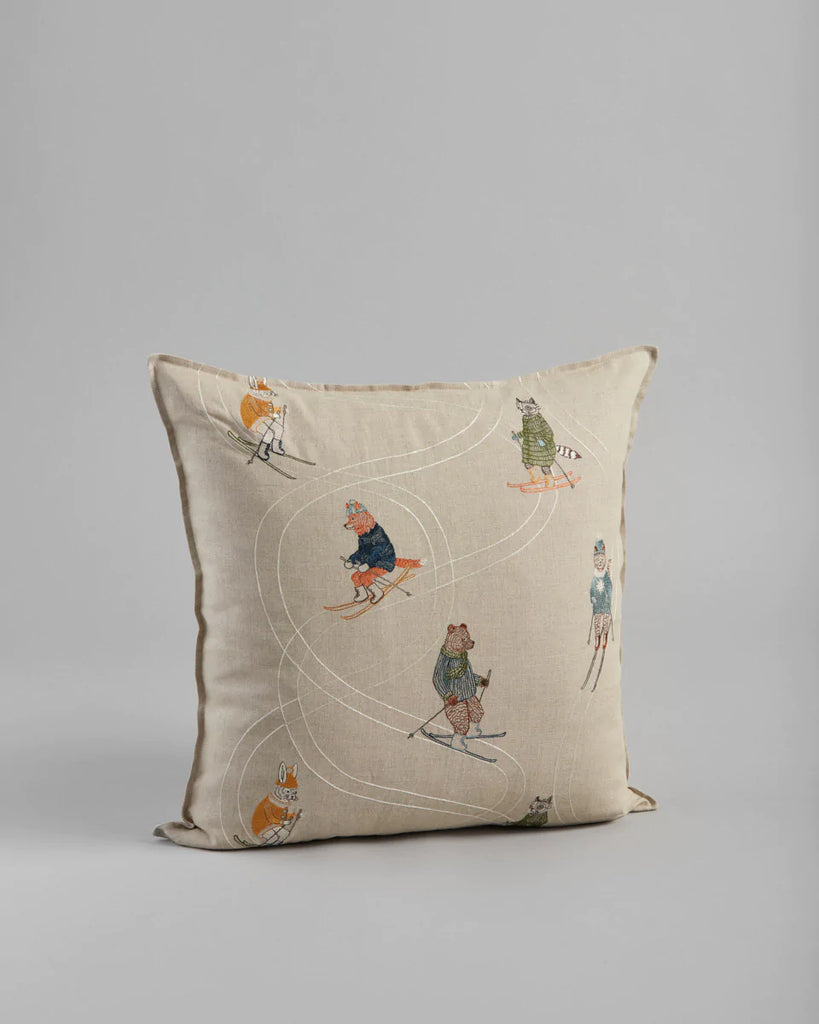 Decorative Coral & Tusk Downhill Skiers Pillow with an embroidery motif of children playing and animals, depicted in a whimsical, circular pattern on a light neutral fabric, set against a plain gray background.