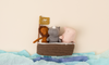 Three Noah's Friends stuffed animal toys—a lion, a rhino, and an elephant—sit inside a small brown basket on a background mimicking water with blue paper.