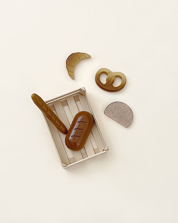 A flat-lay image of Erzi Assorted Baked Goods in Crate including a bread loaf, banana, pretzel, and wooden crate on a light background. The banana and pretzel are outside the crate.