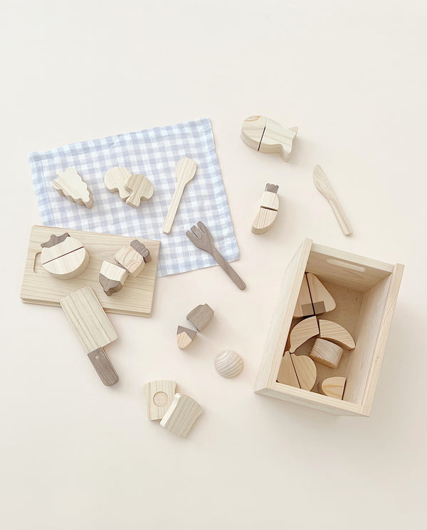 A collection of Hinoki wood toy kitchen utensils and Velcro Wooden Vegetables neatly arranged next to a small wooden crate and a blue and white checkered cloth on a light background.