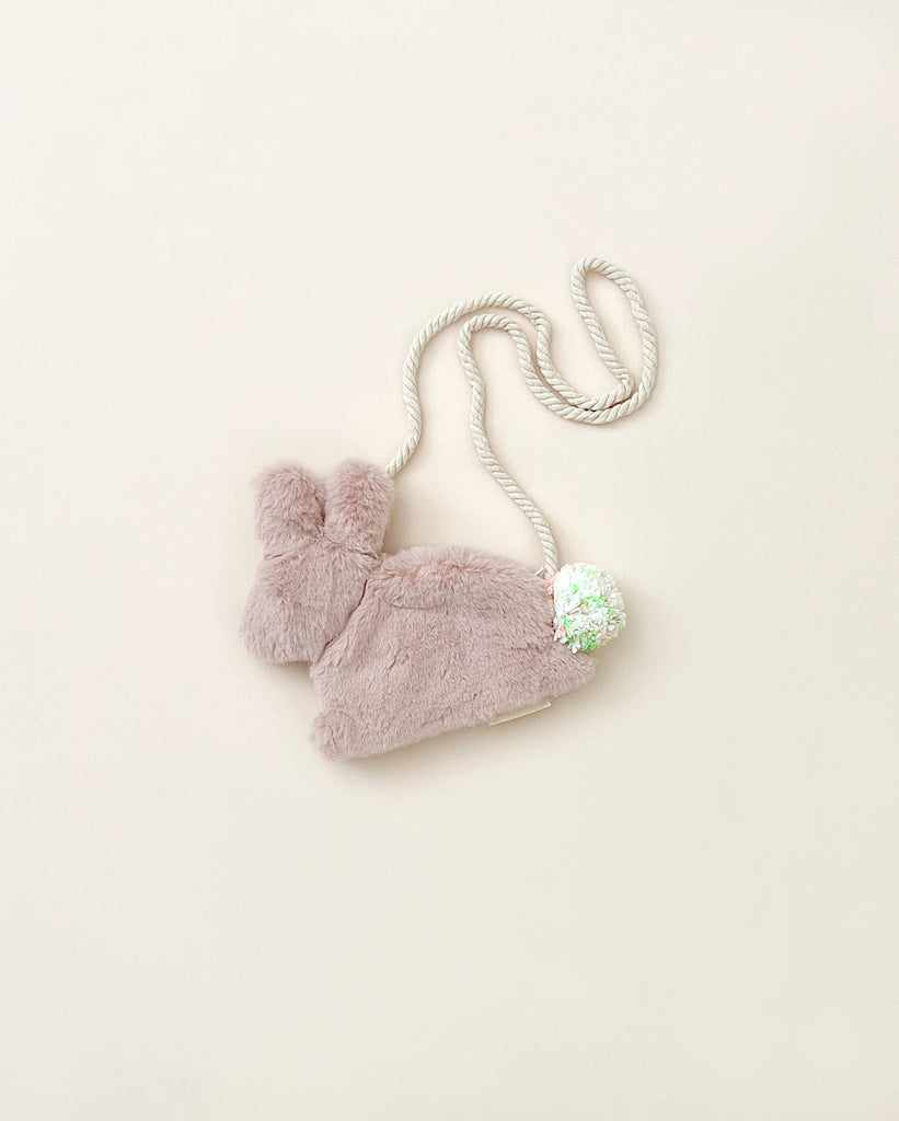 A Meri Meri Plush Bunny Purse, pale pink winter hat with a fluffy pom-pom and a soft, braided tie, displayed against a light beige background.