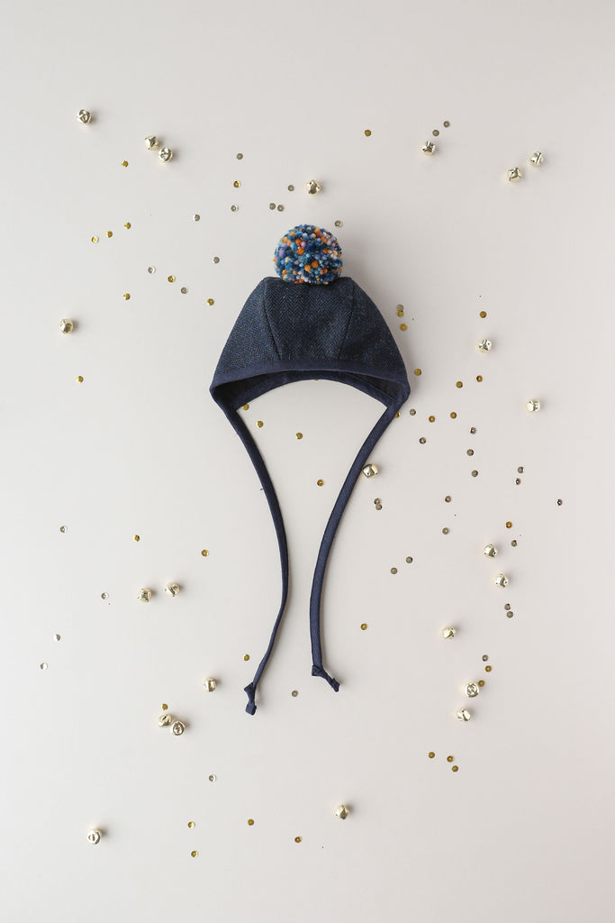 A navy blue Briar Baby Eon Pom Bonnet with ear flaps and a multicolored pom-pom on top, surrounded by small decorative beads scattered on a light background.