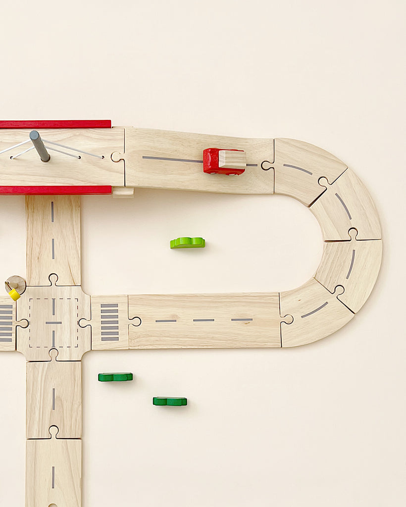 Wooden Road System Deluxe shaped like the letter 'd' with a red and yellow train car, and scattered green pieces on a light background, crafted from reclaimed rubberwood.