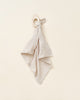 A soft, off-white Organic Muslin Lovey with a grid texture is tied to a wooden teething ring, set against a plain light beige background.