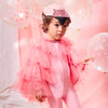 A young child in a Meri Meri Flamingo Costume adorned with pink tulle wings, stands among pink balloons and star decorations, looking thoughtful.